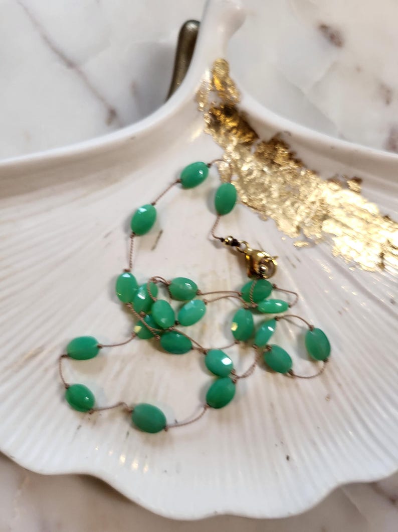 May include: A delicate necklace featuring faceted, oval-shaped green beads strung on a thin, gold-colored chain. The necklace is displayed on a white, shell-shaped dish with gold leaf accents. The clasp is gold-toned.