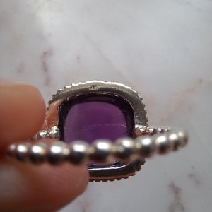 May include: A silver ring with a large, square, deep purple gemstone. The gemstone is surrounded by a halo of small, clear stones. The band of the ring has a beaded design.