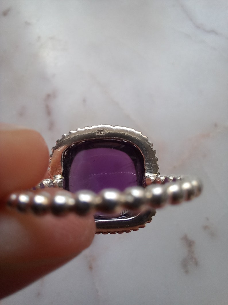 May include: A silver ring with a large, square, deep purple gemstone. The gemstone is surrounded by a halo of small, clear stones. The band of the ring has a beaded design.