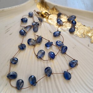 Puede incluir: Un collar delicado con piedras preciosas de cianita azul en forma de l&aacute;grima facetada, ensartadas en un cord&oacute;n fino de color marr&oacute;n. El collar incluye un peque&ntilde;o cierre redondo de metal dorado en la parte superior que une los extremos.