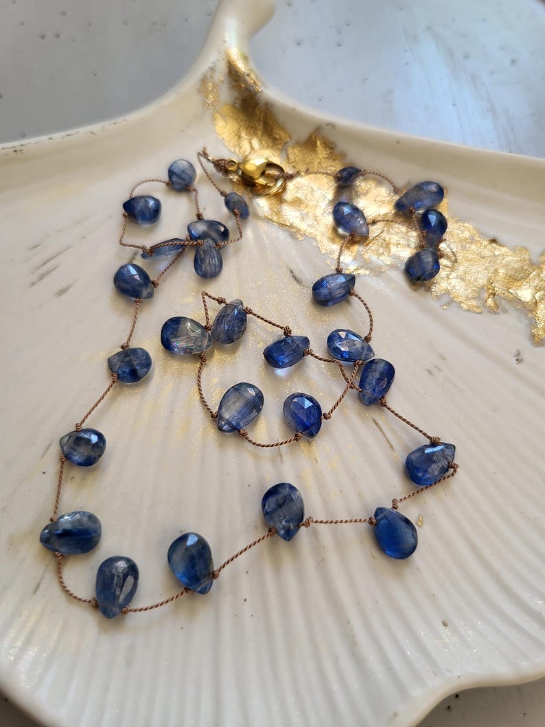 Puede incluir: Un collar delicado con piedras preciosas de cianita azul en forma de l&aacute;grima facetada, ensartadas en un cord&oacute;n fino de color marr&oacute;n. El collar incluye un peque&ntilde;o cierre redondo de metal dorado en la parte superior que une los extremos.