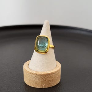 May include: A gold ring with a square blue and green gemstone. The ring is displayed on a wooden ring holder.
