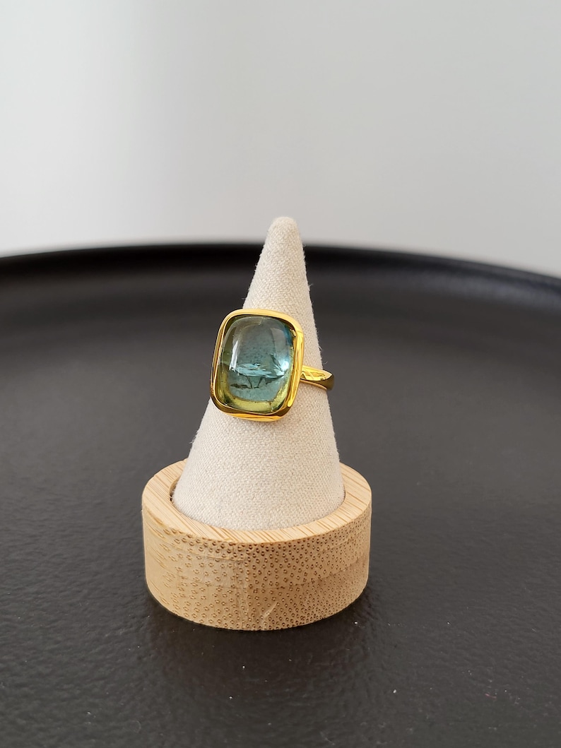 May include: A gold ring with a square blue and green gemstone. The ring is displayed on a wooden ring holder.