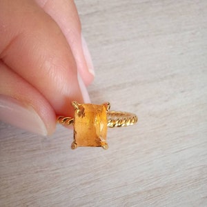 Puede incluir: Un anillo de oro con una piedra preciosa rectangular de color naranja. El anillo tiene una banda trenzada y cuatro garras que aseguran la piedra. La piedra preciosa es de color ámbar cálido.