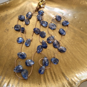 Puede incluir: Un collar con piedras preciosas de cianita azul en forma de l&aacute;grima facetada, ensartadas en un cord&oacute;n fino de color marr&oacute;n claro. El collar incluye un peque&ntilde;o cierre de metal dorado en la parte superior, con las piedras espaciadas a lo largo del cord&oacute;n.