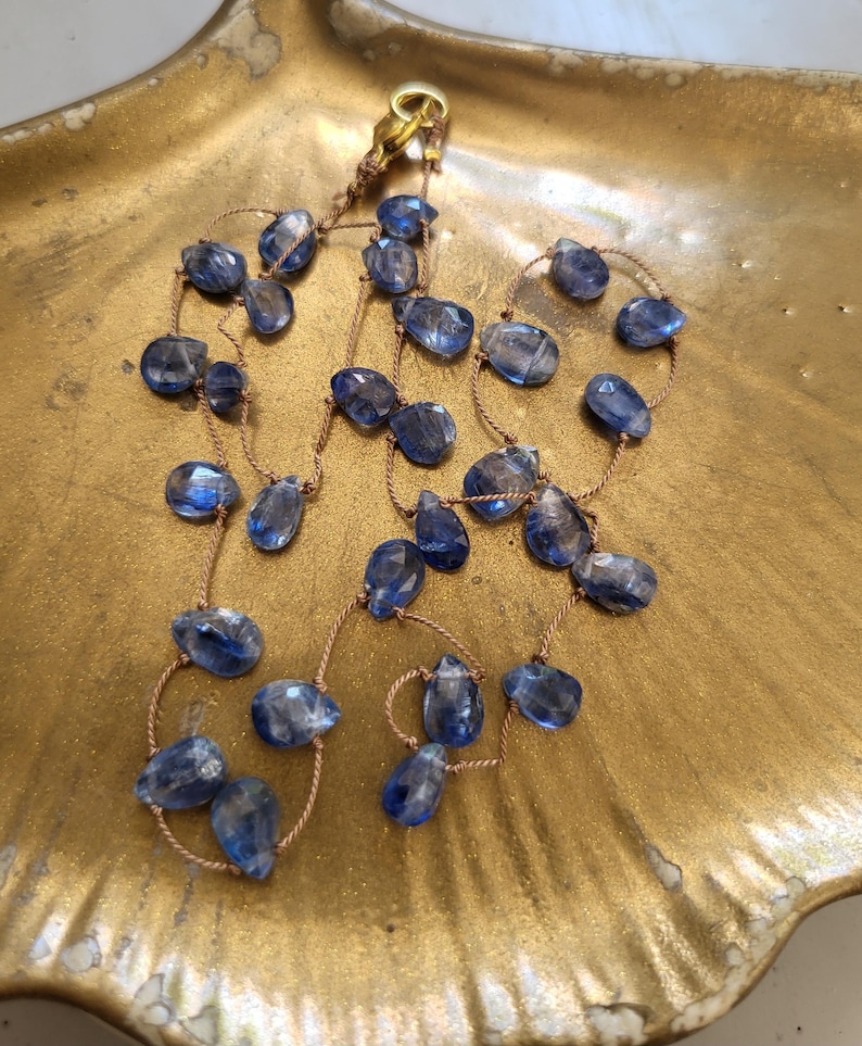 Puede incluir: Un collar con piedras preciosas de cianita azul en forma de l&aacute;grima facetada, ensartadas en un cord&oacute;n fino de color marr&oacute;n claro. El collar incluye un peque&ntilde;o cierre de metal dorado en la parte superior, con las piedras espaciadas a lo largo del cord&oacute;n.