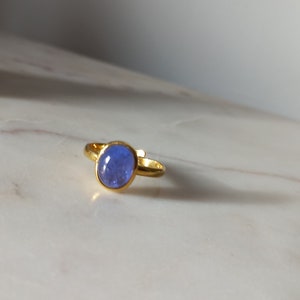 K&ouml;nnte beinhalten: Ein goldener Ring mit einem blauen ovalen Edelstein. Der Ring liegt auf einer wei&szlig;en Marmorfl&auml;che.
