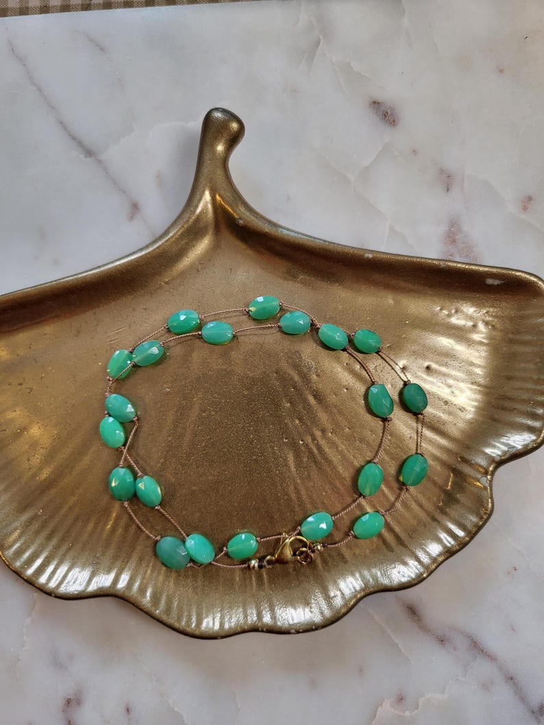 May include: A double-strand necklace featuring oval, faceted, turquoise-colored beads. The beads are strung on a delicate, gold-toned chain. The necklace is displayed on a gold-colored, leaf-shaped dish.