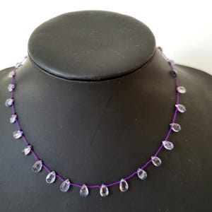 May include: A delicate necklace featuring clear, teardrop-shaped beads strung on a vibrant purple cord. The beads are evenly spaced, creating a minimalist and elegant design. The necklace is displayed on a black mannequin.