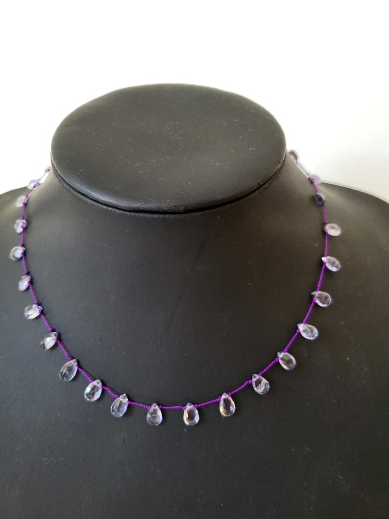 May include: A delicate necklace featuring clear, teardrop-shaped beads strung on a vibrant purple cord. The beads are evenly spaced, creating a minimalist and elegant design. The necklace is displayed on a black mannequin.