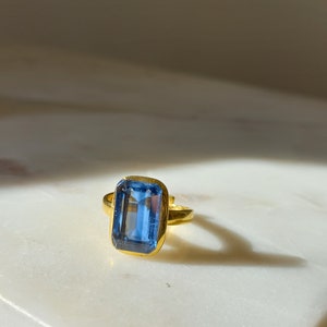 May include: A gold ring featuring a large, rectangular, light blue gemstone. The ring's band and setting are gold-colored, creating a striking contrast with the blue stone. The ring is displayed on a white surface.