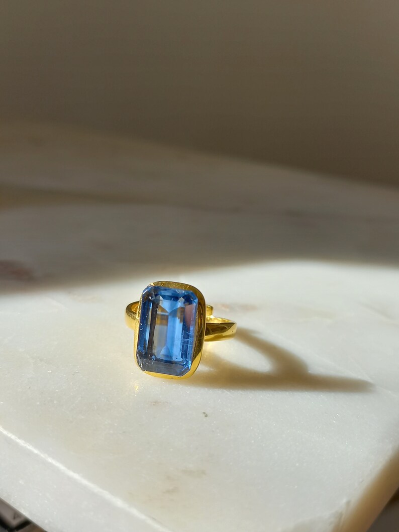 May include: A gold ring featuring a large, rectangular, light blue gemstone. The ring's band and setting are gold-colored, creating a striking contrast with the blue stone. The ring is displayed on a white surface.