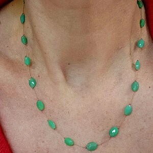 May include: A delicate necklace featuring oval-shaped, light green gemstones strung on a thin, gold-colored chain. The gemstones are evenly spaced, creating a simple yet elegant design. The necklace is shown against a skin tone background, highlighting the jewelry.