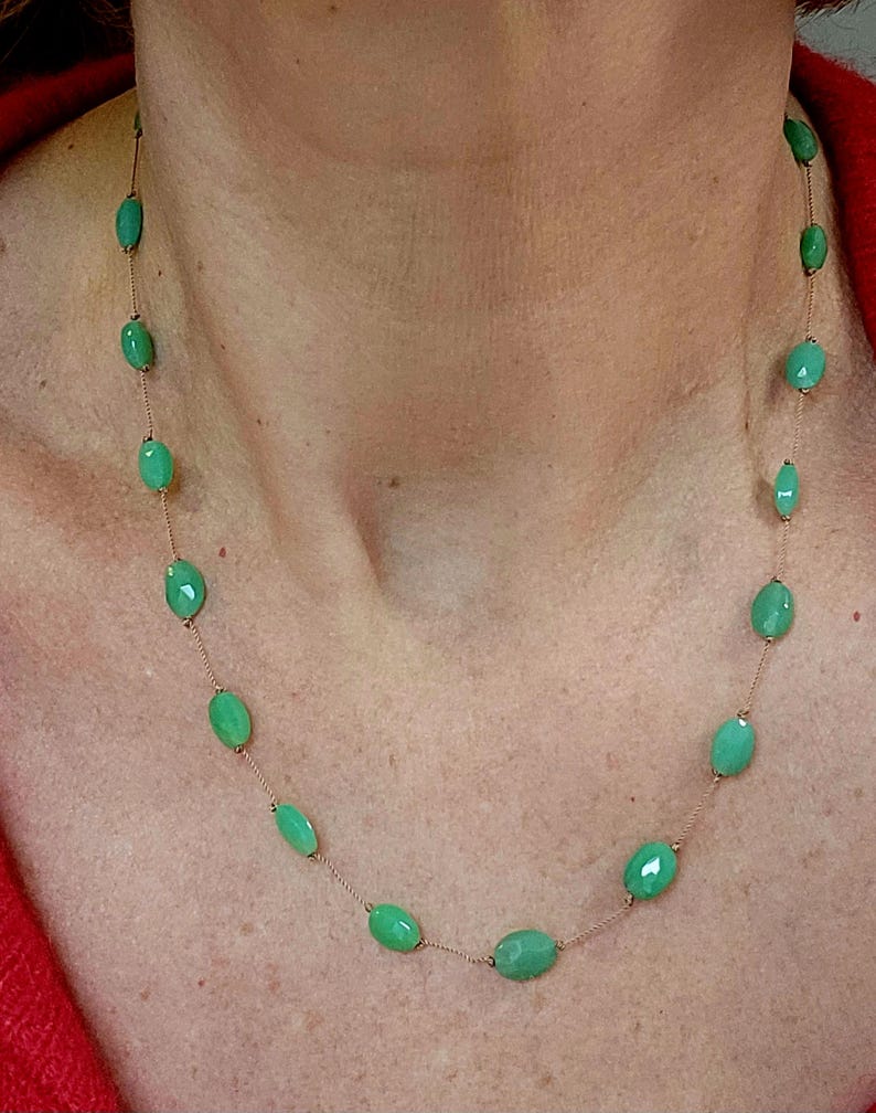 May include: A delicate necklace featuring oval-shaped, light green gemstones strung on a thin, gold-colored chain. The gemstones are evenly spaced, creating a simple yet elegant design. The necklace is shown against a skin tone background, highlighting the jewelry.