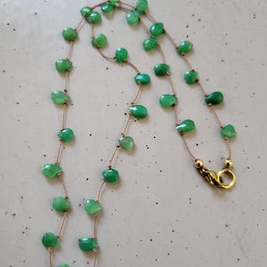 May include: A delicate necklace featuring faceted, light green gemstone beads strung on a thin, tan cord. The necklace has a gold-colored clasp and is displayed against a neutral background. The beads are irregularly shaped and spaced.