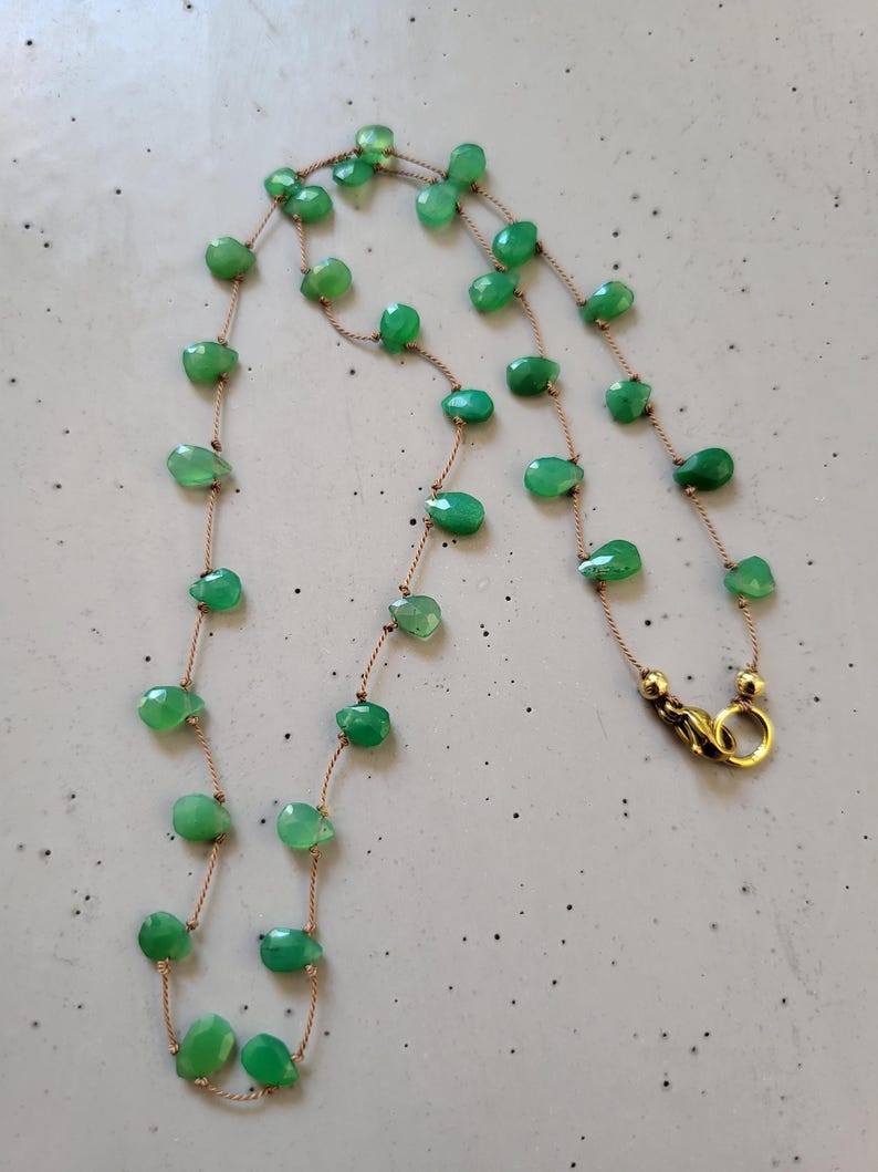 May include: A delicate necklace featuring faceted, light green gemstone beads strung on a thin, tan cord. The necklace has a gold-colored clasp and is displayed against a neutral background. The beads are irregularly shaped and spaced.