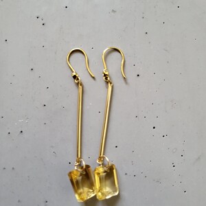 May include: A pair of gold-tone drop earrings featuring a long, slender metal bar suspended from a hook wire. At the base of each bar hangs a rectangular, faceted yellow gemstone that catches the light, creating a clear and bright appearance.