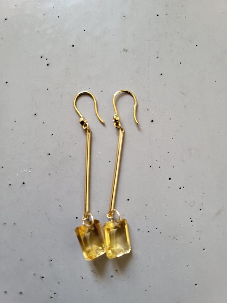 May include: A pair of gold-tone drop earrings featuring a long, slender metal bar suspended from a hook wire. At the base of each bar hangs a rectangular, faceted yellow gemstone that catches the light, creating a clear and bright appearance.