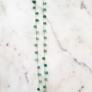 May include: A delicate necklace featuring small, teardrop-shaped green gemstones strung on a thin, brown cord. The necklace has a gold-colored clasp and is displayed against a white and gray marble surface. The gemstones are evenly spaced.