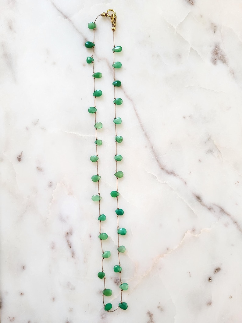 May include: A delicate necklace featuring small, teardrop-shaped green gemstones strung on a thin, brown cord. The necklace has a gold-colored clasp and is displayed against a white and gray marble surface. The gemstones are evenly spaced.