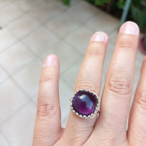 May include: A silver ring featuring a large, square-cut, deep purple gemstone. The gemstone is surrounded by a decorative, scalloped setting. The ring is shown on a finger against a blurred background.