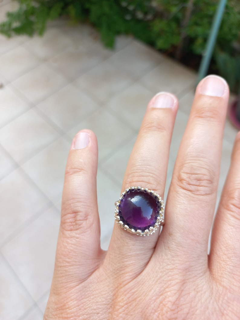 May include: A silver ring featuring a large, square-cut, deep purple gemstone. The gemstone is surrounded by a decorative, scalloped setting. The ring is shown on a finger against a blurred background.