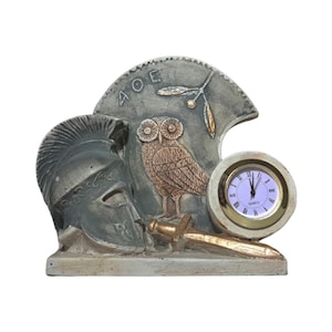 May include: A decorative clock with a gray and gold finish. The clock features a sculpted owl, a Spartan helmet, and a sword. The clock face is white with black Roman numerals and gold hands. The clock is labeled "QUARTZ" and has the Greek word "ΑΘΕ" on the top.
