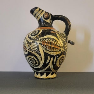 Puede incluir: Una pequeña jarra de cerámica negra y blanca con un asa marrón y diseños geométricos intrincados. La jarra tiene un cuello estrecho y un cuerpo redondeado.