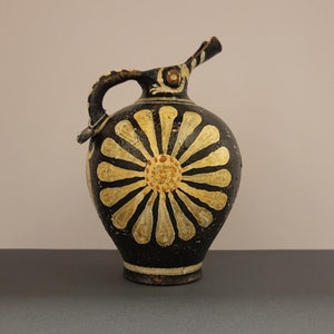 Puede incluir: Jarra de cerámica negra con diseño floral. La jarra presenta una gran flor amarilla y blanca en la parte delantera, con asa y pico. Probablemente sea una pieza antigua o vintage.