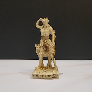 May include: A white ceramic statue of Artemis, the Greek goddess of the hunt, riding a deer. She is holding a bow and arrow in her right hand. The statue is on a base with the word "ARTEMIS" inscribed on it.