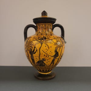 May include: A black and yellow ceramic amphora with a lid. The amphora is decorated with a black and white scene of people harvesting fruit from a tree.