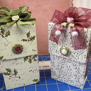 May include: Two decorative gift boxes. One box features a green bow and a red jewel button. The other box has a burgundy bow and a pearl button. Both boxes are decorated with holly and berry patterns, perfect for holiday gifting.