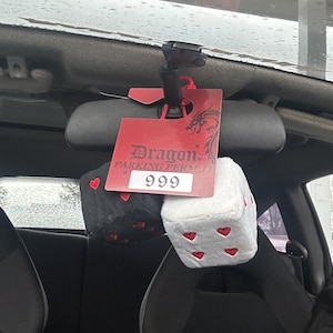 May include: A red parking permit with the word "Dragon" and the number "999" is hanging from a car's rearview mirror. Below the permit are two fuzzy dice, one black with red hearts and one white with red hearts.