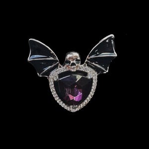 May include: A silver bat brooch featuring a purple crystal and a skull at its centre. The bat wings are black and the crystal is surrounded by clear rhinestones.