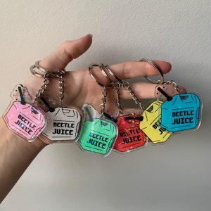 May include: A collection of six acrylic keychains in various colors, each featuring a drink design with the text "BEETLE JUICE". The keychains have silver metal rings and chains, with glitter accents.