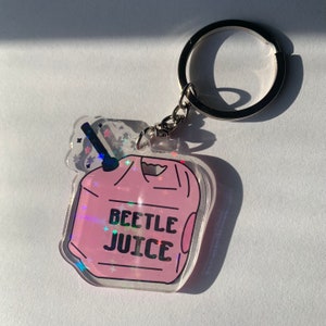 May include: A clear acrylic keychain with a pink design featuring the words "BEETLE JUICE" in black lettering. The keychain has a silver key ring and chain. The design has a holographic effect.