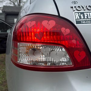 May include: A silver car with a red rear light. The rear light has several broken heart stickers on it. The stickers are red and white. The car has a sticker on the back window that says "R's Fire, Cambridge, 740-432".