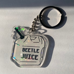 May include: A clear acrylic keychain with a holographic design. The keychain features a cartoon-style image of a beverage with the words "BEETLE JUICE" in black. The keychain is attached to a silver chain and keyring.
