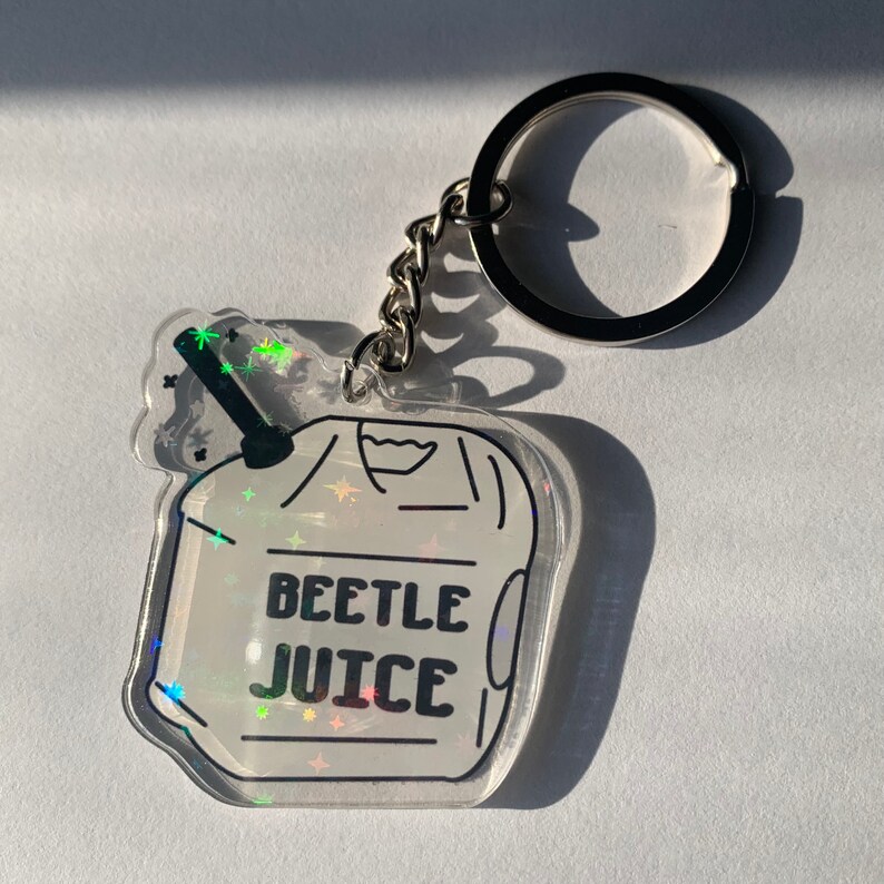 May include: A clear acrylic keychain with a holographic design. The keychain features a cartoon-style image of a beverage with the words "BEETLE JUICE" in black. The keychain is attached to a silver chain and keyring.