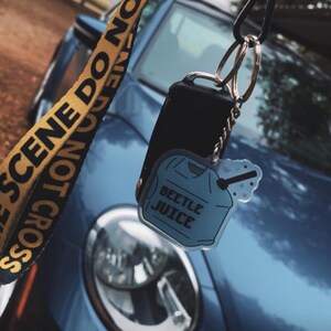 May include: A blue car key fob with a "Beetle Juice" keychain. The keychain is a light blue shirt shape with the words "Beetle Juice" printed on it. A yellow and black lanyard with the words "SCENE DO NOT CROSS" is also visible.