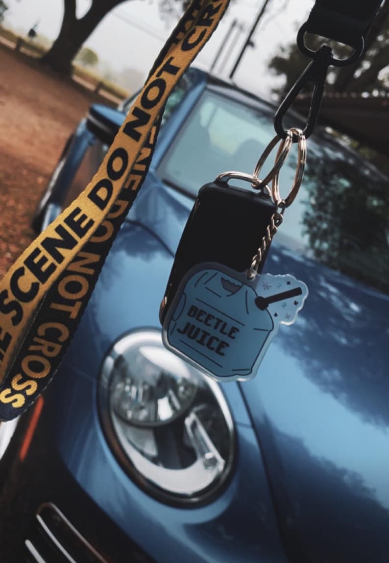 May include: A blue car key fob with a "Beetle Juice" keychain. The keychain is a light blue shirt shape with the words "Beetle Juice" printed on it. A yellow and black lanyard with the words "SCENE DO NOT CROSS" is also visible.