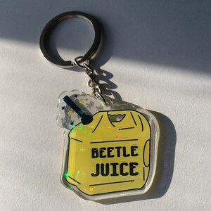 May include: A clear acrylic keychain with a yellow design featuring the words "BEETLE JUICE" in black. The keychain has a silver keyring and chain. The design includes a black straw and star accents.