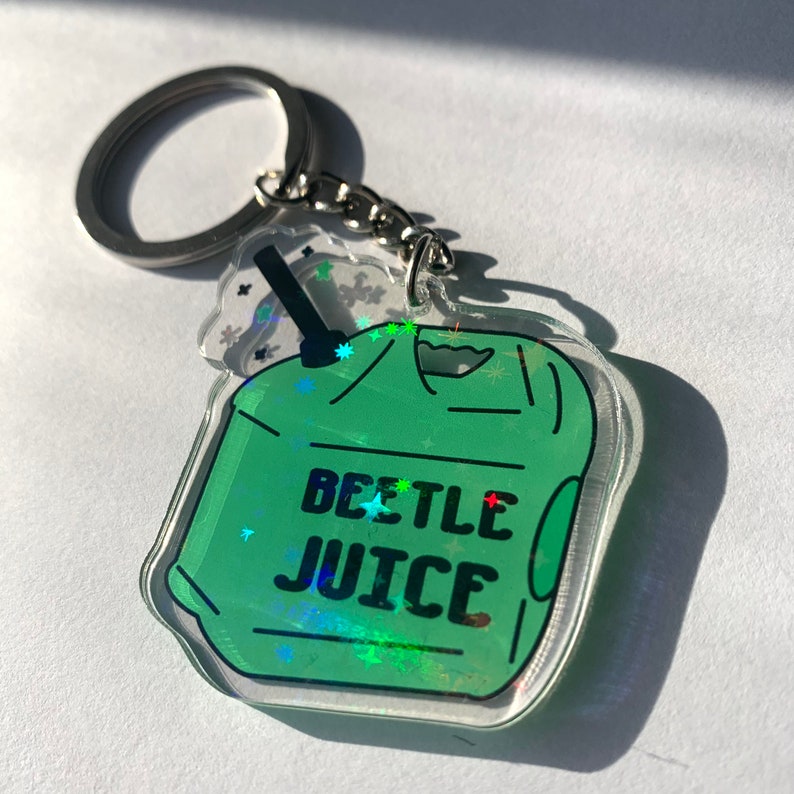 May include: A clear acrylic keychain with a green cartoon beverage design and the words "BEETLE JUICE" in black. The keychain has a silver metal ring and chain.