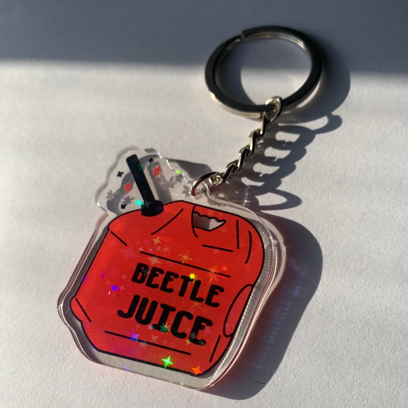 May include: A clear acrylic keychain with a red design that says "BEETLEJUICE" in black letters. The keychain has a silver key ring and chain. The design has a holographic effect with glittery stars.