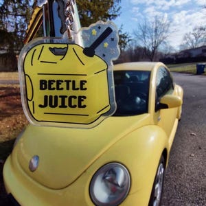 May include: A yellow keychain with the text "BEETLE JUICE" in black letters, hanging in front of a yellow Volkswagen Beetle car. The keychain has a yellow gas can design with a black nozzle and a star-shaped accent.
