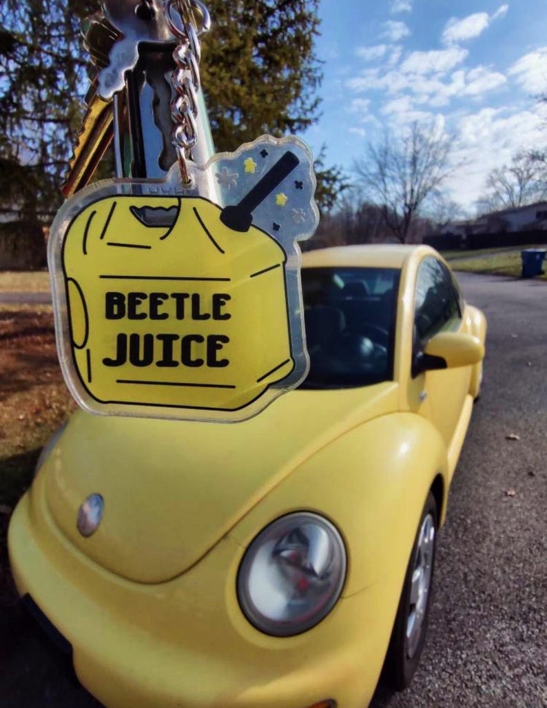 May include: A yellow keychain with the text "BEETLE JUICE" in black letters, hanging in front of a yellow Volkswagen Beetle car. The keychain has a yellow gas can design with a black nozzle and a star-shaped accent.