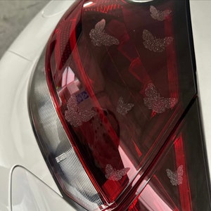 May include: A white car with a red taillight. The taillight has a decorative design of multiple silver glitter butterflies.