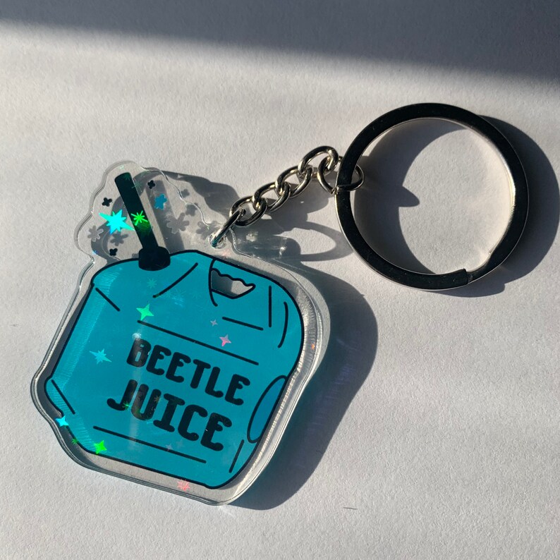 May include: A clear acrylic keychain with a blue design that says "BEETLE JUICE". The keychain has a silver chain and a large silver key ring. The design includes stars and a drink illustration.