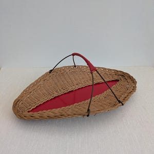 May include: Oval wicker serving tray with a red interior. The tray has a woven brown exterior and a red handle. The handle is wrapped in red material. The tray has a black metal frame.