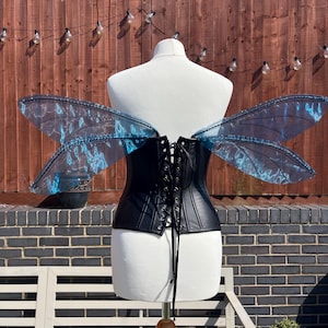 May include: Black leather corset with a lace-up back, worn on a white mannequin. The mannequin is wearing a pair of large, blue, iridescent fairy wings.